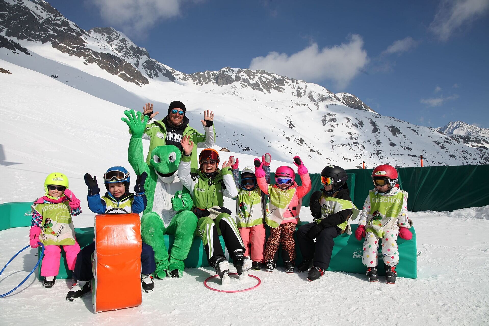 Eine Gruppe von Kindern und Erwachsenen in Skikleidung posiert und winkt vor verschneiten Bergen; eine Person trägt dabei ein grünes Frosch-Maskottchenkostüm.