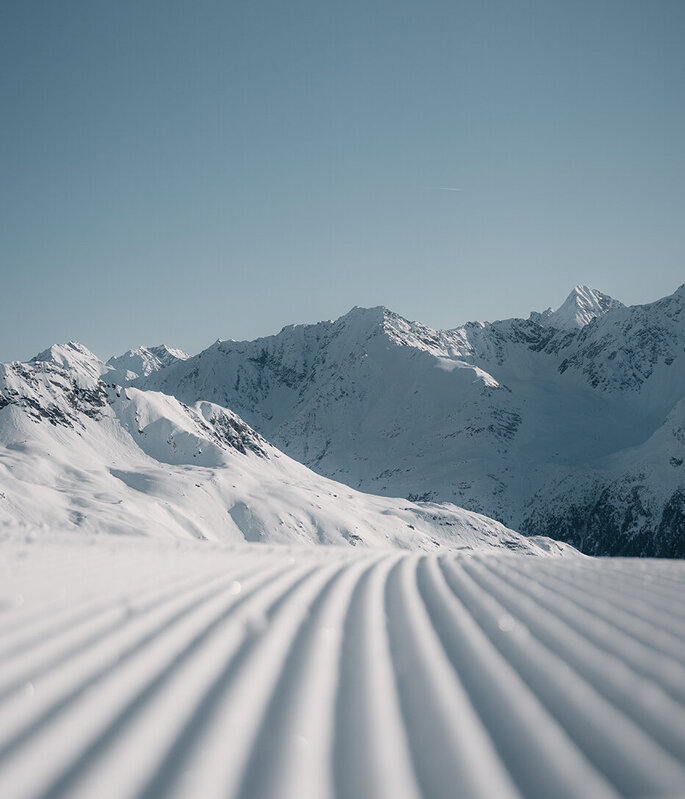 Eine frisch präparierte Skipiste mit parallelen Spuren im Schnee, die unter einem strahlend blauen Himmel auf die fernen, schneebedeckten Berge zuführen.