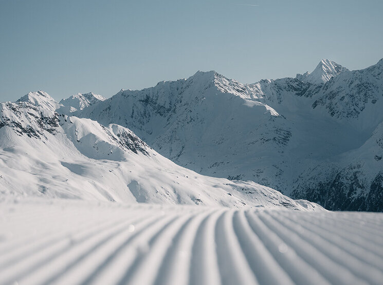Eine frisch präparierte Skipiste mit parallelen Spuren im Schnee, die unter einem strahlend blauen Himmel auf die fernen, schneebedeckten Berge zuführen.
