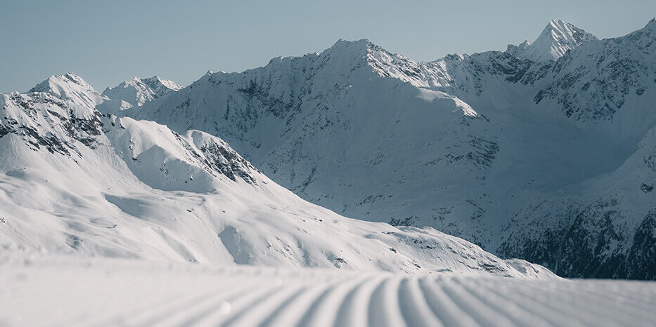 Eine frisch präparierte Skipiste mit parallelen Spuren im Schnee, die unter einem strahlend blauen Himmel auf die fernen, schneebedeckten Berge zuführen.