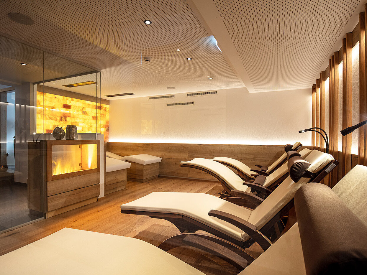 Ein moderner Wellness-Ruheraum mit mehreren geschwungenen Liegestühlen, sanfter Beleuchtung, Holzwänden und einer leuchtenden Salzwand in der Ecke, die eine warme, ruhige Atmosphäre schafft.