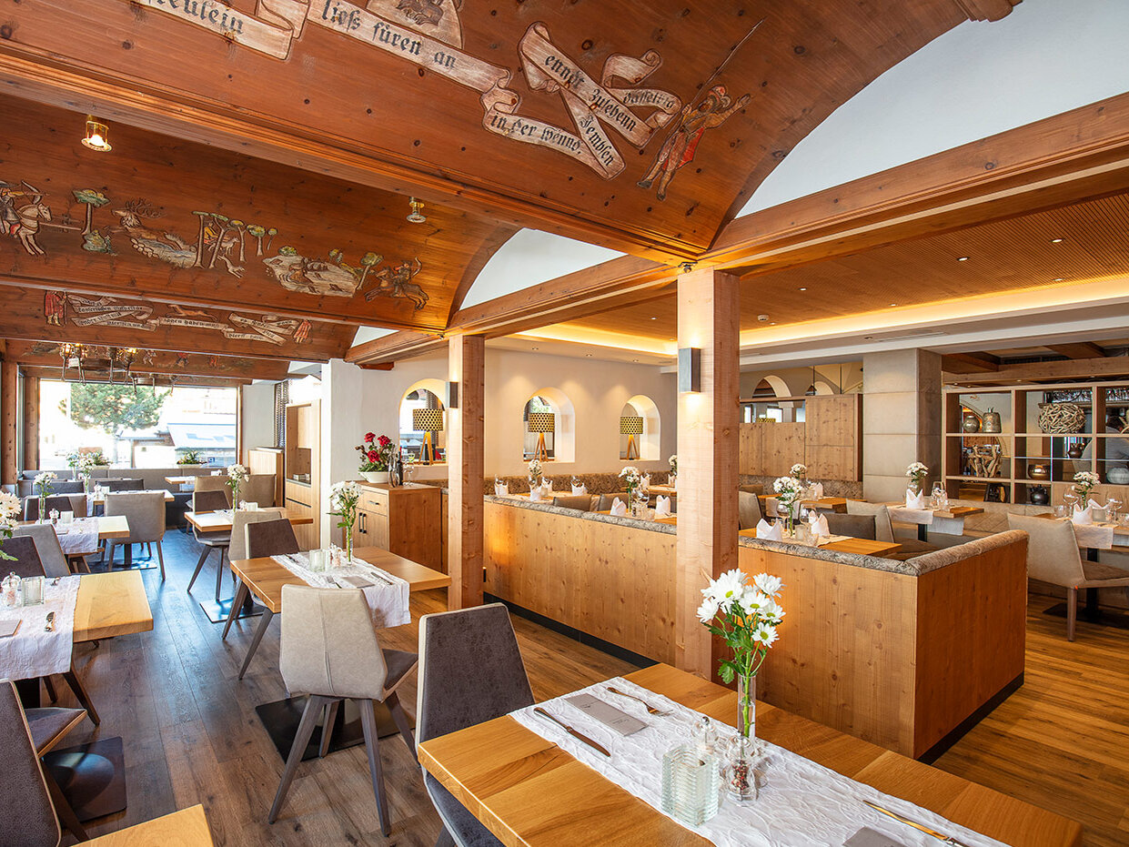 Ein helles, modernes Restaurant mit Holztischen und -stühlen, Blumengestecken und dekorativen Holzdecken. Große Fenster lassen Tageslicht herein und schaffen so eine warme und einladende Atmosphäre.