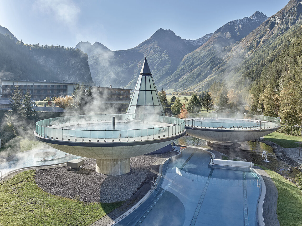 Zwei große, dampfende, runde Außenpools und eine gläserne Pyramidenkonstruktion liegen inmitten von Bergen und Bäumen, verbunden durch einen gewundenen blauen Pool unter strahlend blauem Himmel.