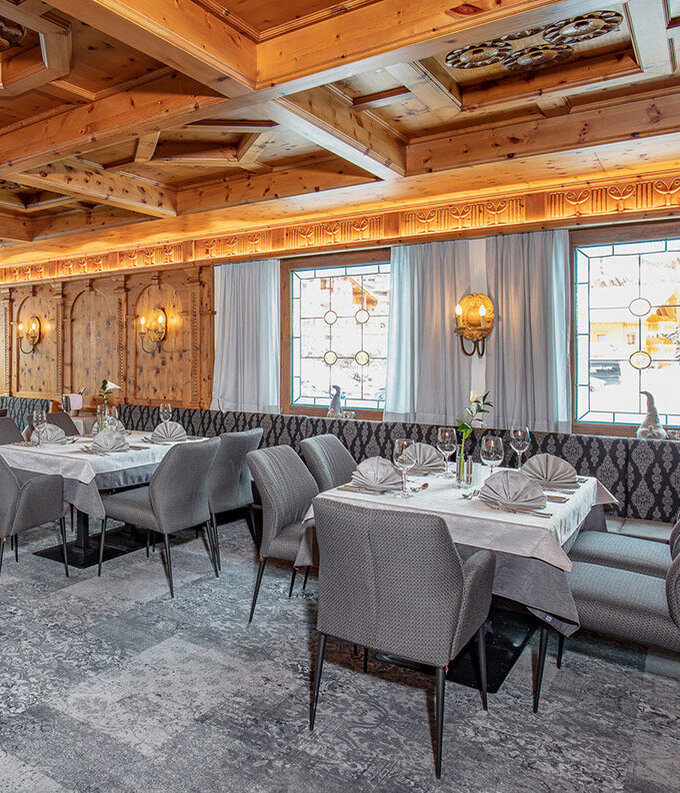 Ein elegantes Restaurant mit gemustertem Teppichboden, grauen Polsterstühlen, weißen Tischdecken und einer holzvertäfelten Decke. Große Fenster lassen Tageslicht herein, und Wandleuchten sorgen für eine warme Atmosphäre. Die Tische sind ordentlich für das Essen gedeckt.