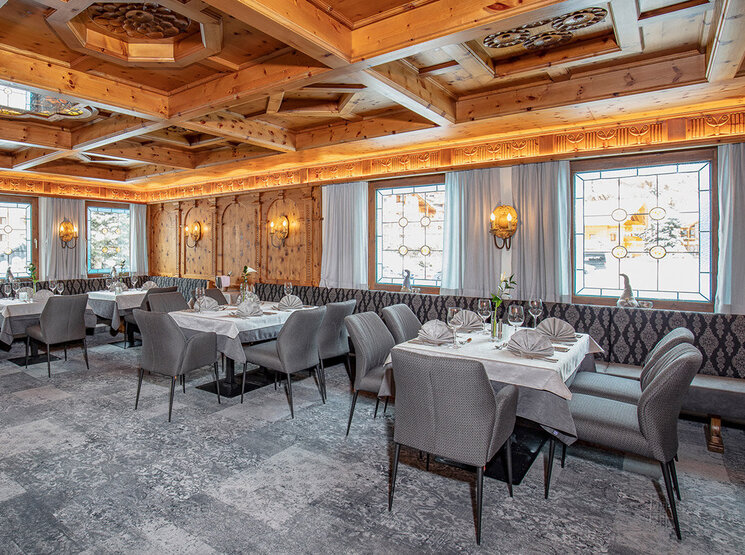 Ein elegantes Restaurant mit gemustertem Teppichboden, grauen Polsterstühlen, weißen Tischdecken und einer holzvertäfelten Decke. Große Fenster lassen Tageslicht herein, und Wandleuchten sorgen für eine warme Atmosphäre. Die Tische sind ordentlich für das Essen gedeckt.