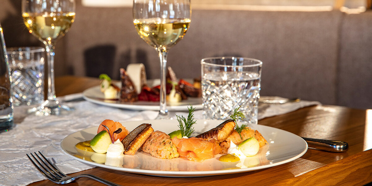  Auf einem Holztisch steht ein Teller mit Gourmet-Lachs, Brotchips und Beilagen; im Hintergrund sind Weingläser, Wassergläser und ein Dessertteller zu sehen.