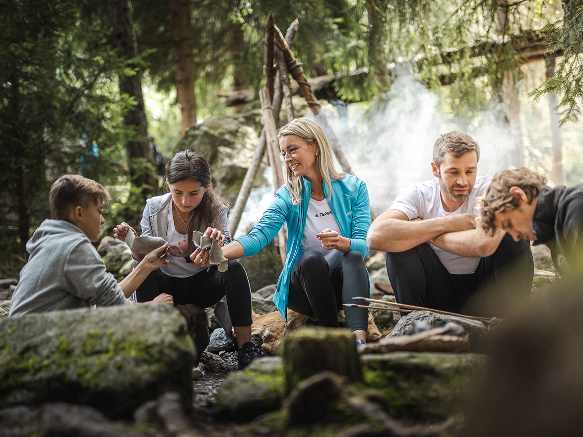 Vier junge Erwachsene sitzen auf Felsen in einem Wald, umgeben von Bäumen. Entdeckung im Ötzi-Dorf.