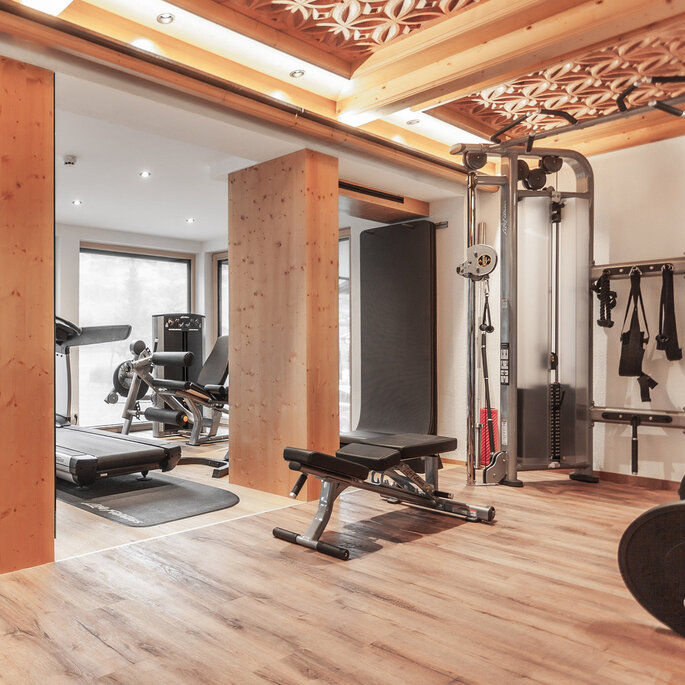 Ein modernes Fitnessstudio mit Holzelementen, ausgestattet mit einem Laufband, einer Hantelbank, einem Krafttrainingsgerät, freien Gewichten und großen Fenstern, durch die Tageslicht hereinströmt.