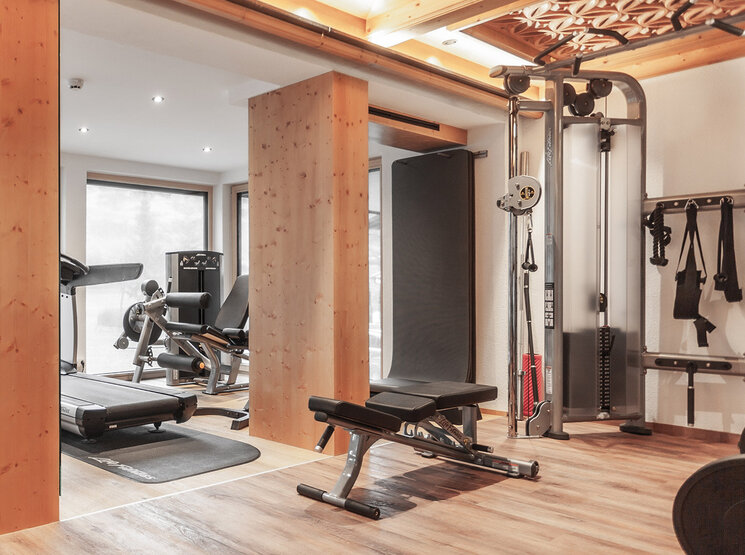 Ein modernes Fitnessstudio mit Holzelementen, ausgestattet mit einem Laufband, einer Hantelbank, einem Krafttrainingsgerät, freien Gewichten und großen Fenstern, durch die Tageslicht hereinströmt.