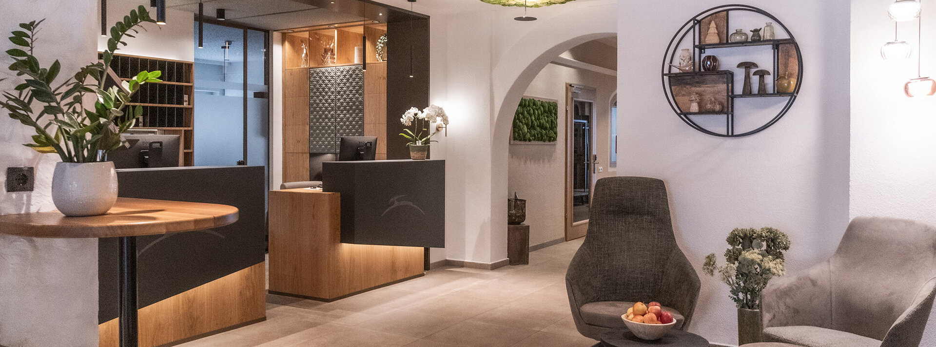 Moderne Hotellobby mit Rezeption, stilvollen Stühlen, rundem Wandregal mit Dekoration, Pflanzen und sanfter Beleuchtung, die eine einladende und gemütliche Atmosphäre schaffen.