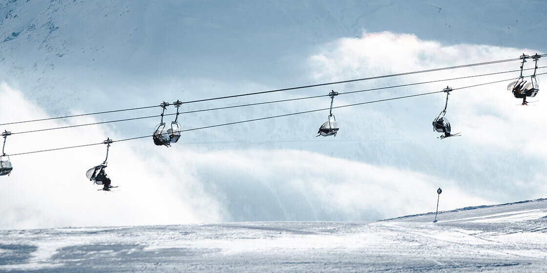 Skifahrer fahren mit einem Sessellift einen verschneiten Berghang hinauf, im Hintergrund ragt unter klarem Himmel ein hoher, schroffer, schneebedeckter Gipfel empor.