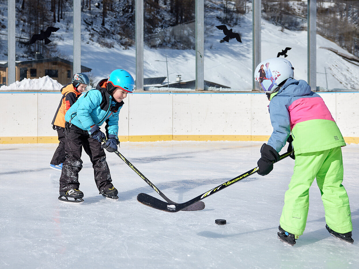 Drei Kinder in Winterkleidung spielen draußen Eishockey. Zwei kämpfen um den Puck, während ein drittes hinter ihnen herläuft. Im Hintergrund sind eine verschneite Landschaft und hohe Fenster zu sehen.