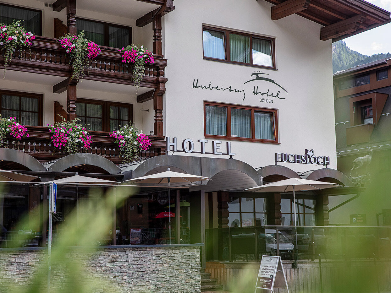 Außenansicht des Hotels mit Balkonen, die mit rosa Blumen geschmückt sind, großen Fenstern und einem Schild mit der Aufschrift „Hotel Hubertus“. Im Vordergrund sind Sitzgelegenheiten im Freien mit Sonnenschirmen sowie eine Steinmauer und Grünpflanzen zu sehen.