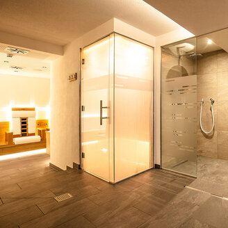 Moderner Wellnessbereich mit einer verglasten Dusche, Aromadampfbad mit Milchglastüren, Fliesenboden und sanfter Stimmungsbeleuchtung. Im Hintergrund ist eine Sauna mit Holzbänken zu sehen.