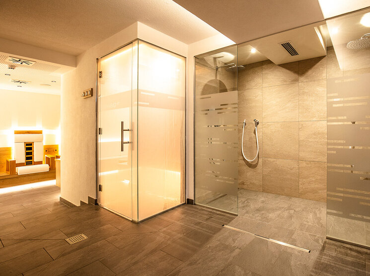 Moderner Wellnessbereich mit einer verglasten Dusche, Aromadampfbad mit Milchglastüren, Fliesenboden und sanfter Stimmungsbeleuchtung. Im Hintergrund ist eine Sauna mit Holzbänken zu sehen.