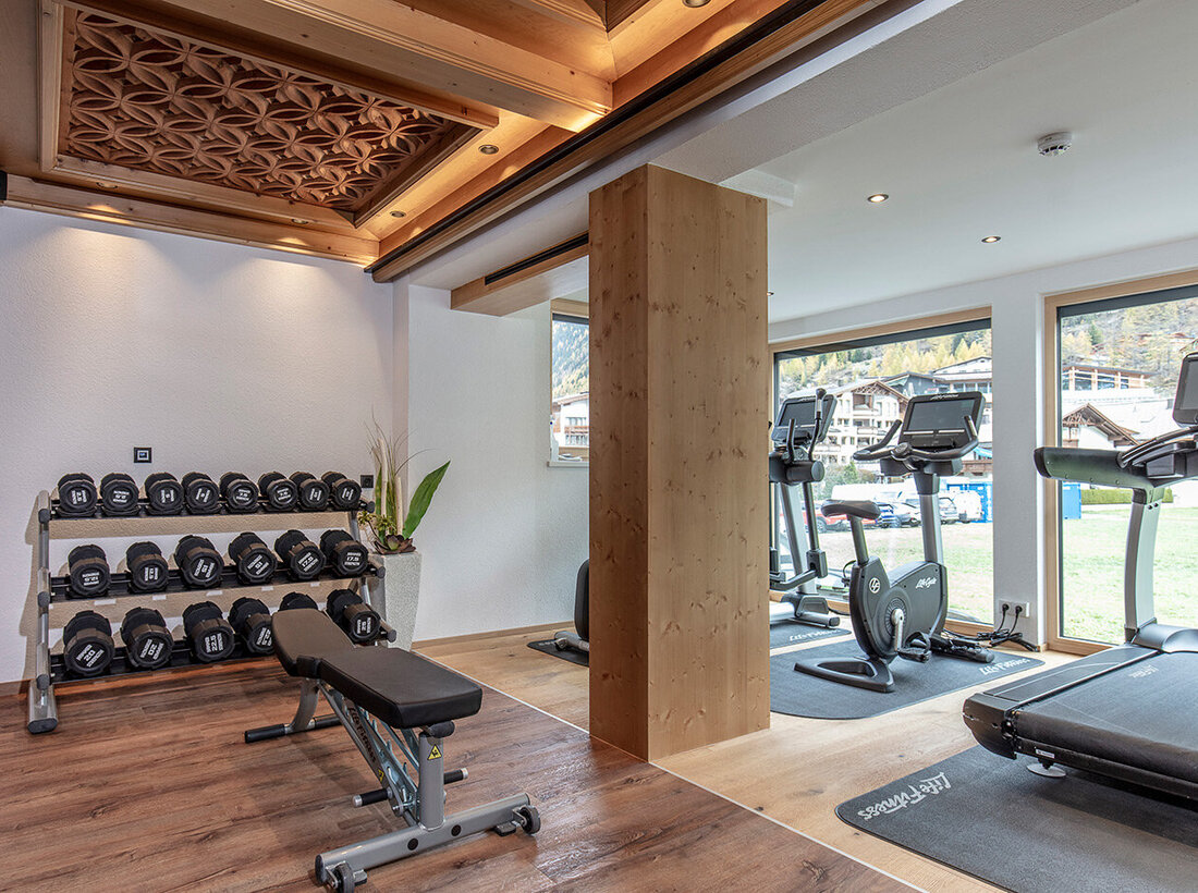 Ein moderner Fitnessraum mit einem Hantelständer, einer Hantelbank, Laufbändern und Heimtrainern vor großen Fenstern; Holzböden und dekorative Deckenpaneele sorgen für eine gemütliche Atmosphäre.