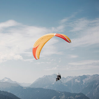 Zwei Menschen, die mit einem gelb-orangefarbenen Gleitschirm über eine Bergkette mit hohen, schneebedeckten Gipfeln gleiten, vor einem blauen Himmel mit vereinzelten Wolken.