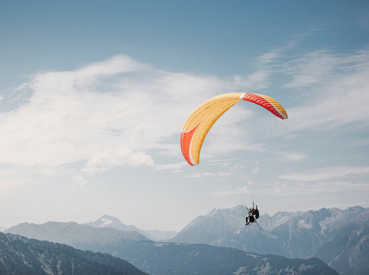 Zwei Menschen, die mit einem gelb-orangefarbenen Gleitschirm über eine Bergkette mit hohen, schneebedeckten Gipfeln gleiten, vor einem blauen Himmel mit vereinzelten Wolken.