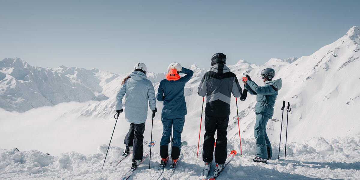 Vier Skifahrer stehen auf einem verschneiten Berghang und blicken auf eine malerische Aussicht auf schneebedeckte Gipfel unter einem klaren Himmel; ihre Skistöcke und ihre Ausrüstung sind zu sehen.