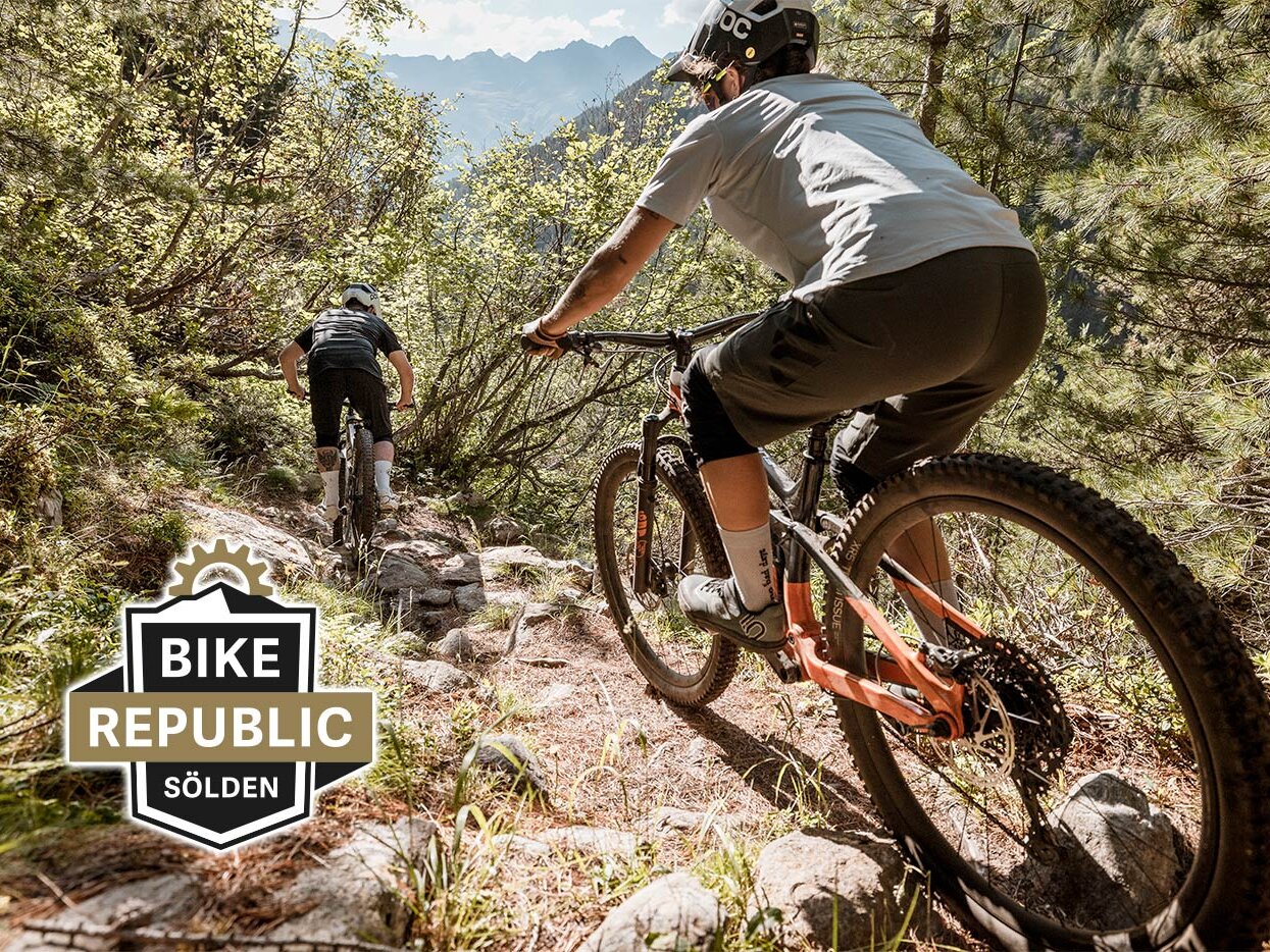 Zwei Mountainbiker fahren im Sonnenlicht einen felsigen Waldweg hinunter, umgeben von Bäumen und Bergen. Im Vordergrund ist das Logo von „Bike Republic Sölden“ zu sehen.