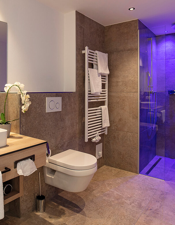 Modernes Badezimmer mit einem Waschbecken auf einem Waschtisch aus Holz, einem WC, einem beheizten Handtuchhalter und einer Glasdusche mit violetter Beleuchtung. Neutrale Farbtöne und eine Orchidee schmücken den Raum.