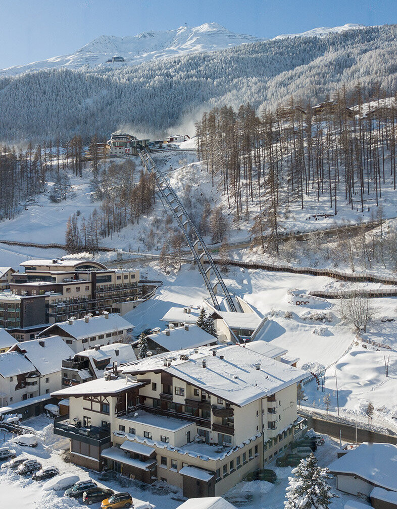 Ein verschneites Bergdorf mit Häusern, geparkten Autos und schneebedeckten Bäumen. Der Zentrum Shuttle führt hinter dem Hotel Hubertus den Hang hinauf, im Hintergrund sind Berge und ein strahlend blauer Himmel zu sehen.