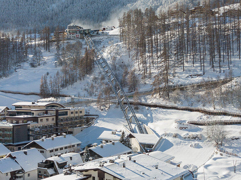  Ein verschneites Bergdorf mit Häusern, geparkten Autos und schneebedeckten Bäumen. Der Zentrum Shuttle führt hinter dem Hotel Hubertus den Hang hinauf, im Hintergrund sind Berge und ein strahlend blauer Himmel zu sehen.