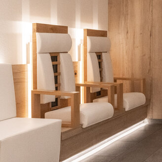 Zwei moderne Infrarot-Holzstühle mit weißen Kissen und Armlehnen stehen nebeneinander in einem minimalistischen, hell beleuchteten Raum mit Holzwänden und Fliesenboden.
