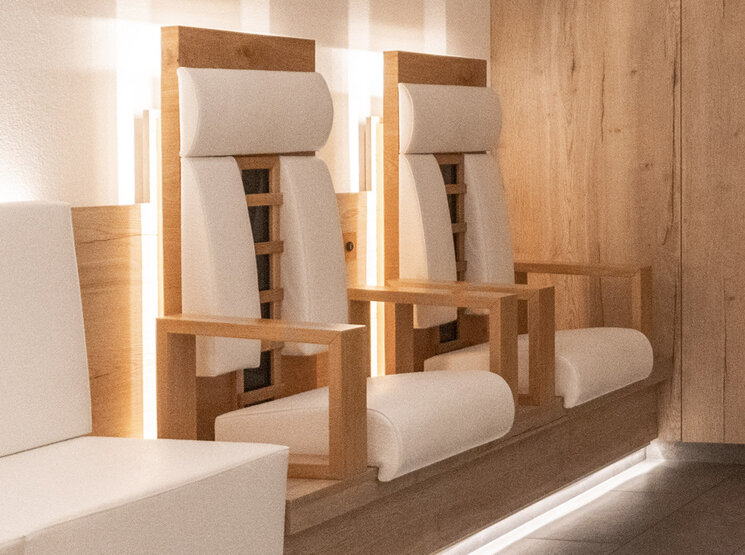 Zwei moderne Infrarot-Holzstühle mit weißen Kissen und Armlehnen stehen nebeneinander in einem minimalistischen, hell beleuchteten Raum mit Holzwänden und Fliesenboden.