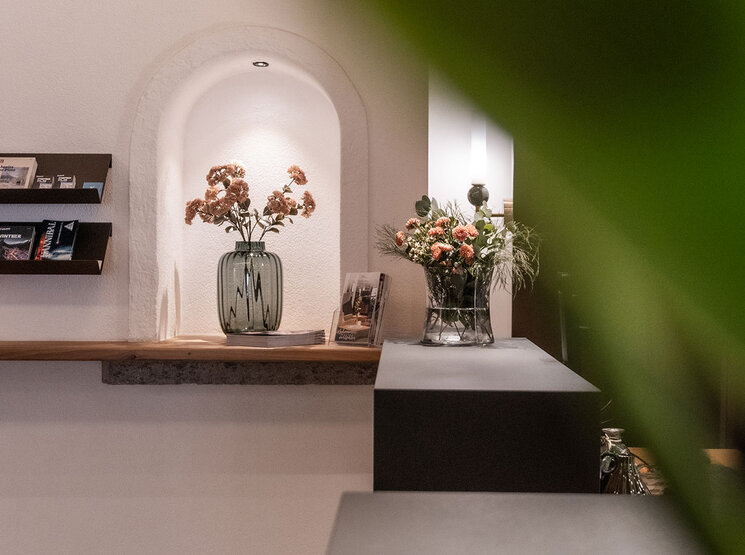 In einem sanft beleuchteten Innenraum stehen in einer Nische eine Vase mit Trockenblumen, Prospekte auf Ausstellungsregalen und eine weitere Vase mit frischen Blumen. Ein grünes Blatt verdeckt teilweise die rechte Seite des Bildes.