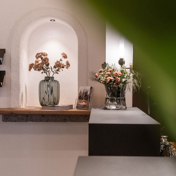 In einem sanft beleuchteten Innenraum stehen in einer Nische eine Vase mit Trockenblumen, Prospekte auf Ausstellungsregalen und eine weitere Vase mit frischen Blumen. Ein grünes Blatt verdeckt teilweise die rechte Seite des Bildes.