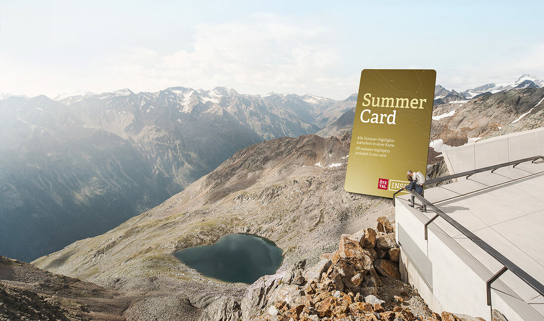 Eine Person sitzt auf einer modernen Terrasse mit Blick auf eine Berglandschaft, unter der sich ein See erstreckt. Auf einer großen Grafik steht „Summer Card“. Im Hintergrund erstrecken sich schneebedeckte Gipfel unter einem strahlend blauen Himmel.