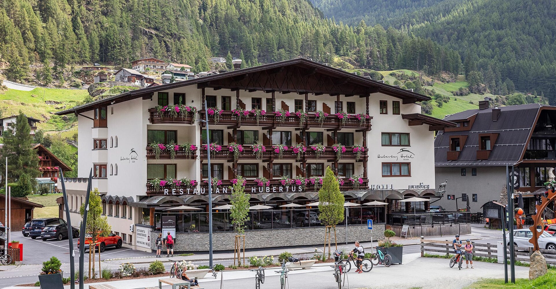 Ein großes Hotel mit Balkonen, die mit rosa Blumen geschmückt sind, umgeben von Bergen und viel Grün. In der Nähe stehen Fahrräder, und im Vordergrund sitzen Menschen auf Bänken.