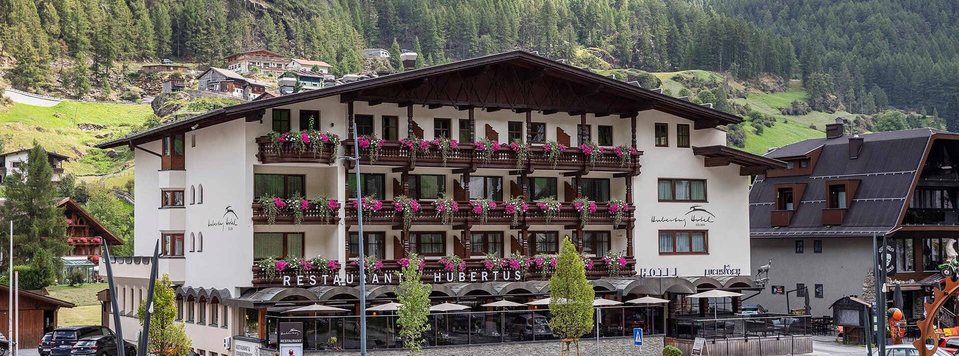 Ein großes Hotel mit Balkonen, die mit rosa Blumen geschmückt sind, umgeben von Bergen und viel Grün. In der Nähe stehen Fahrräder, und im Vordergrund sitzen Menschen auf Bänken.