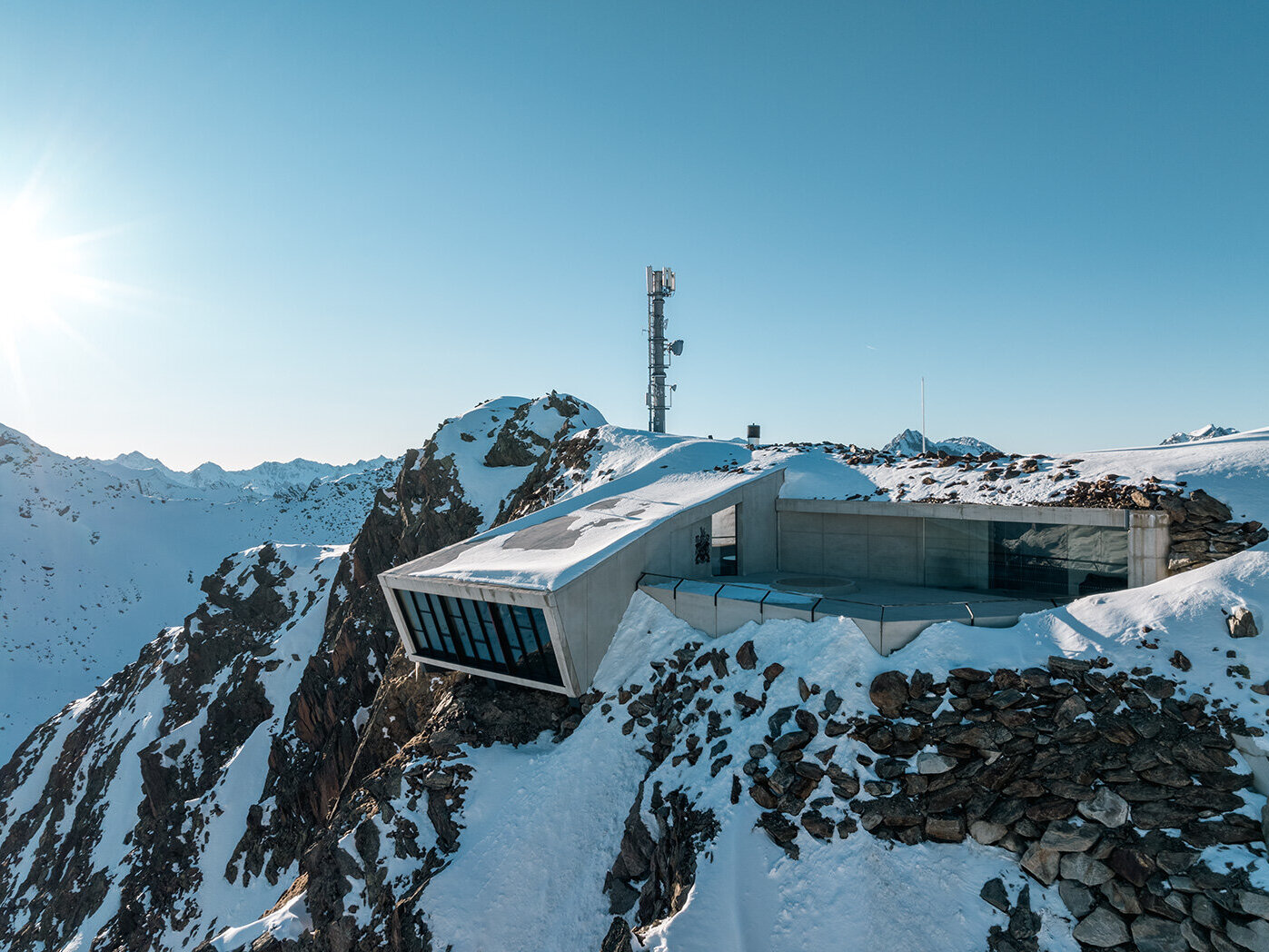 Ein modernes Gebäude aus Glas und Beton, das 007 Elements, das auf einem schneebedeckten Bergkamm thront, mit einem Funkturm in der Nähe und schneebedeckten Gipfeln in der Ferne unter einem strahlend blauen Himmel.