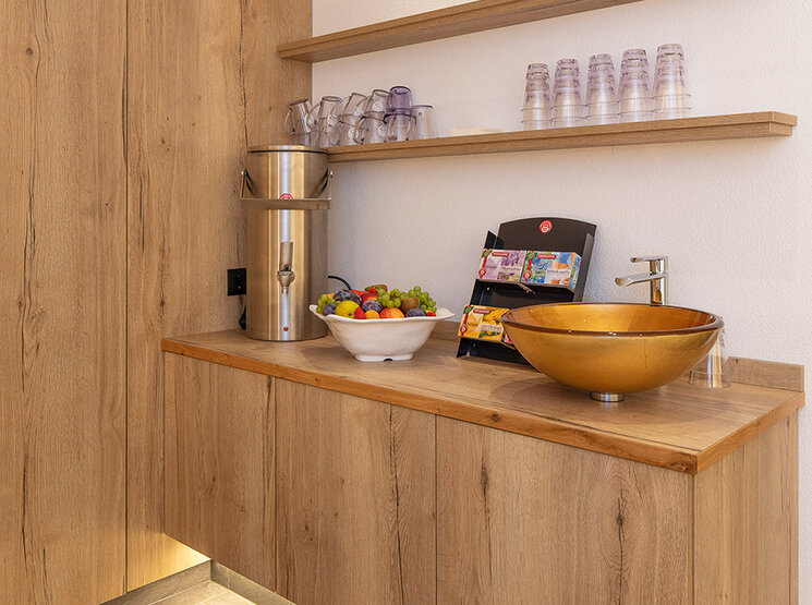 Moderne Teebar mit Holzschränken, einer goldenen Spüle, einer Obstschale, einer Snackauslage, Gläsern, Flaschen und einer Unterbauleuchte. Auf zwei Holzregalen stehen Tassen und Getränke. Das Design ist minimalistisch und hell.