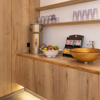 Moderne Teebar mit Holzschränken, einer goldenen Spüle, einer Obstschale, einer Snackauslage, Gläsern, Flaschen und einer Unterbauleuchte. Auf zwei Holzregalen stehen Tassen und Getränke. Das Design ist minimalistisch und hell.