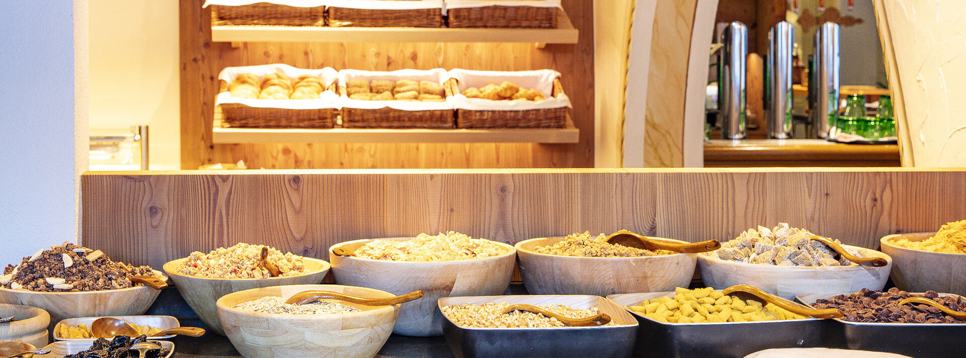 Ein Frühstücksbuffet mit verschiedenen Müsli- und Getreidesorten in Holzschalen, im Hintergrund stehen auf Regalen Körbe mit Brot und Brötchen.