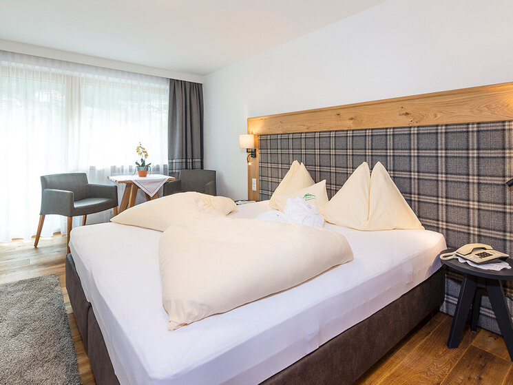 Ein helles Hotelzimmer mit einem Doppelbett, einem karierten Kopfteil, zwei beigen Stühlen an einem runden Tisch neben einem großen Fenster mit transparenten Vorhängen, einem Holzboden und einem Nachttisch mit einem Telefon.