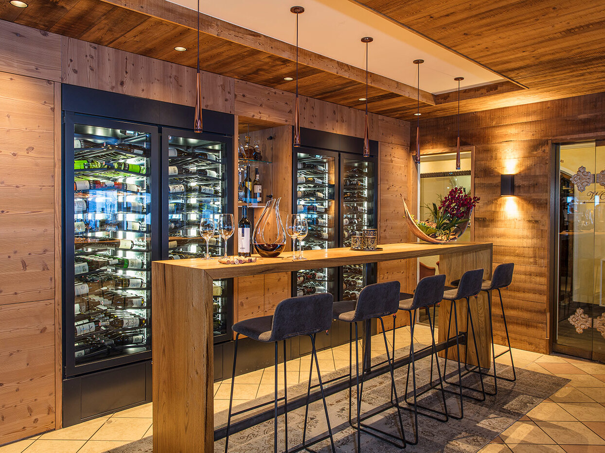 Eine gemütliche Weinbar mit einer Holztheke, vier schwarzen Barhockern und mit Flaschen gefüllten Weinkühlschränken mit Glasfront. Warme Beleuchtung und holzvertäfelte Wände schaffen eine einladende, gehobene Atmosphäre.