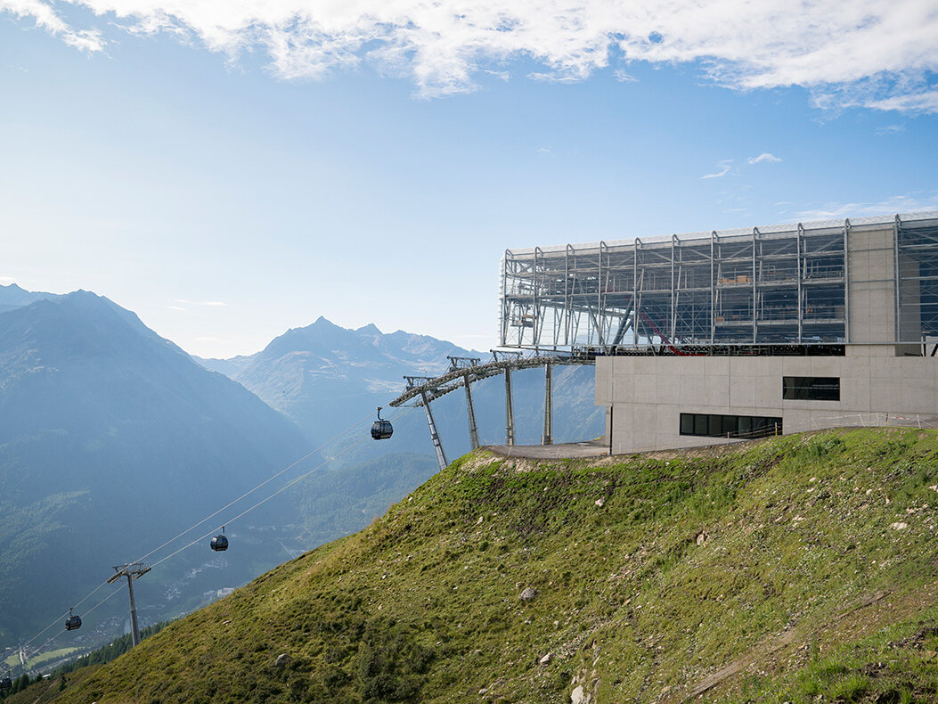 An einem grasbewachsenen Berghang befindet sich eine moderne Seilbahnstation, von der aus Seilbahnen hin- und herfahren. Im Hintergrund sind Berggipfel und ein teilweise bewölkter Himmel zu sehen.