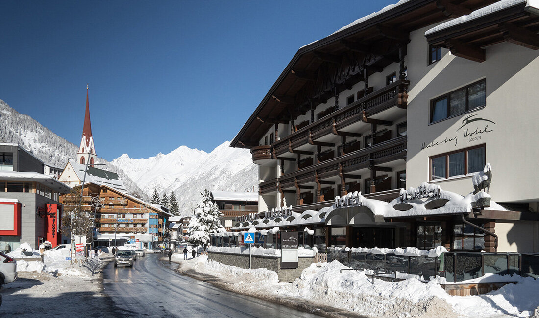 Eine verschneite Straße in einem Bergdorf mit einem Kirchturm, geparkten Autos, Fußgängern und Gebäuden im Chalet-Stil vor dem Hintergrund schneebedeckter Berge unter einem strahlend blauen Himmel.