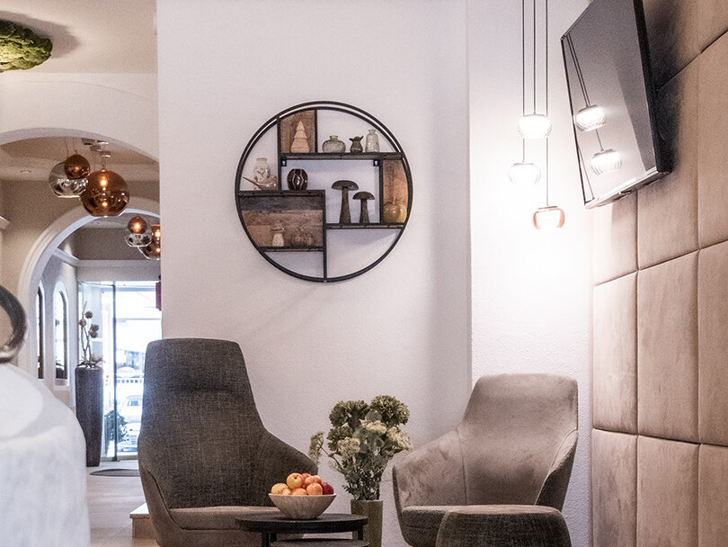 Moderne Lounge mit zwei grauen Stühlen, einem kleinen runden Tisch mit einer Obstschale, Wandbildern und einem an der Wand montierten Fernseher. Der Raum ist hell und besticht durch eine neutrale Farbpalette sowie dekorative Beleuchtung.