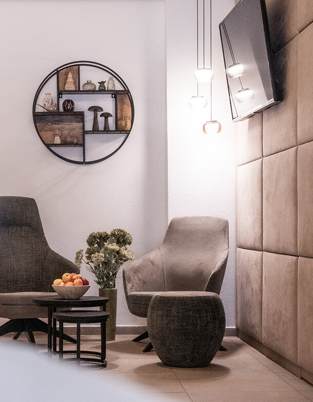 Moderne Lounge mit zwei grauen Stühlen, einem kleinen runden Tisch mit einer Obstschale, Wandbildern und einem an der Wand montierten Fernseher. Der Raum ist hell und besticht durch eine neutrale Farbpalette sowie dekorative Beleuchtung.