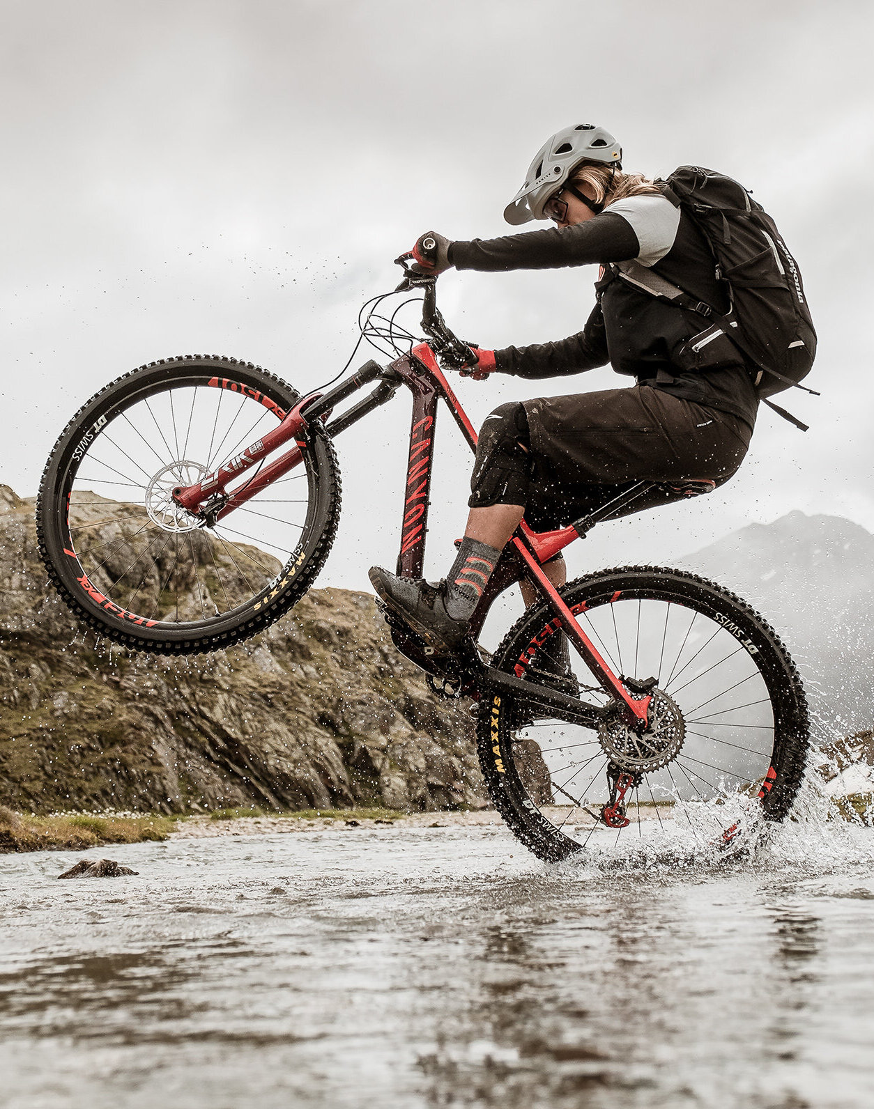 Eine Person mit Helm und Rucksack fährt auf einem roten Mountainbike im Wheelie durch einen seichten, felsigen Bach, während im Hintergrund Hügel und bewölkter Himmel zu sehen sind.
