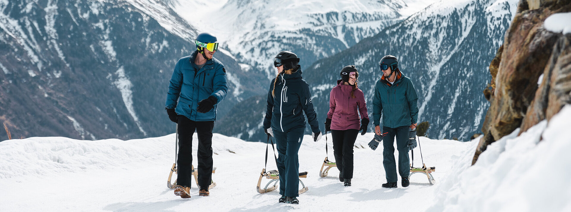 Vier Menschen in Winterkleidung gehen mit Schlitten auf einem verschneiten Bergpfad spazieren, umgeben von schneebedeckten Gipfeln und Bäumen, und genießen einen Wintertag in der freien Natur.