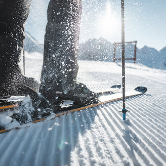 Nahaufnahme der Skischuhe einer Person, die auf einer präparierten, verschneiten Piste in die Skier eingeklickt sind, mit einem Skistock und aufwirbelndem Schnee. Im Hintergrund sind Berge und Sonnenlicht zu sehen.