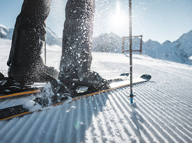 Nahaufnahme der Skischuhe einer Person, die auf einer präparierten, verschneiten Piste in die Skier eingeklickt sind, mit einem Skistock und aufwirbelndem Schnee. Im Hintergrund sind Berge und Sonnenlicht zu sehen.