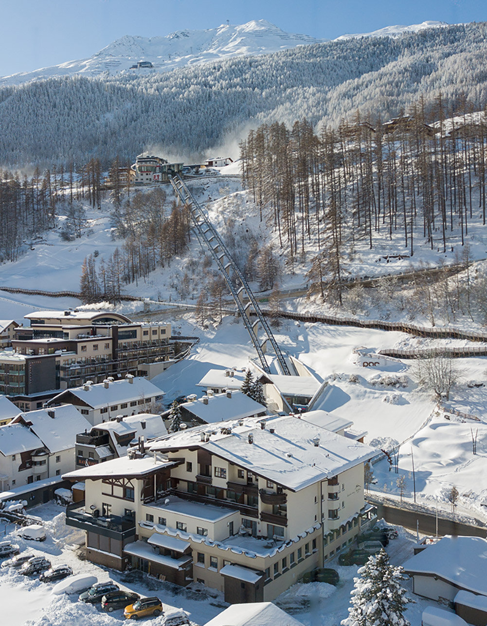 Ein verschneites Bergdorf mit Häusern, geparkten Autos und schneebedeckten Bäumen. Der Zentrum Shuttle führt hinter dem Hotel Hubertus den Hang hinauf, im Hintergrund sind Berge und ein strahlend blauer Himmel zu sehen.