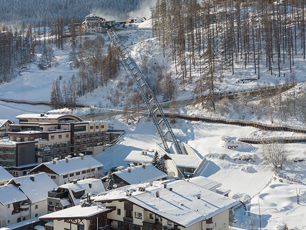Ein verschneites Bergdorf mit Häusern, geparkten Autos und schneebedeckten Bäumen. Der Zentrum Shuttle führt hinter dem Hotel Hubertus den Hang hinauf, im Hintergrund sind Berge und ein strahlend blauer Himmel zu sehen.